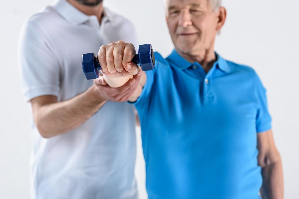 részleges kilátás nyílik a rehabilitációs terapeuta segíti vezető férfi súlyzó elszigetelt szürke gyakorlása - Fotó, kép