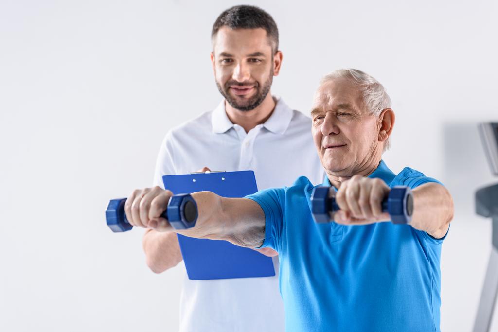 portrét rehabilitačním pracovníkem s Poznámkový blok pomáhat starší muž cvičení s činkami na šedém pozadí - Fotografie, Obrázek