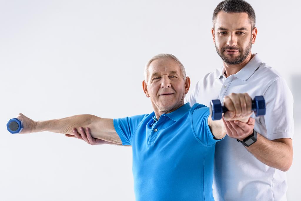 muotokuva kuntoutusterapeutista, joka avustaa harmaalla pohjalla käsipainoilla harjoittelevaa vanhempaa miestä
 - Valokuva, kuva