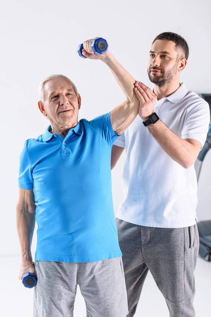 портрет реабилитационного терапевта, помогающего старшему мужчине тренироваться с гантелями на сером фоне
 - Фото, изображение