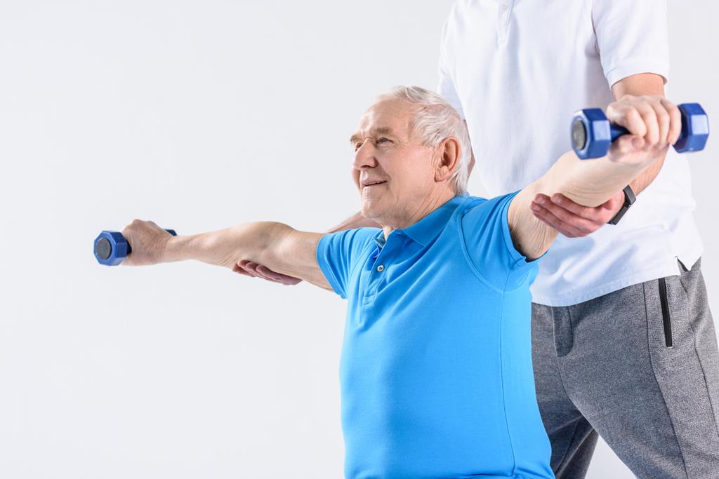 částečný pohled rehabilitační pracovník pomáhat starší muž cvičení s činkami na šedém pozadí - Fotografie, Obrázek