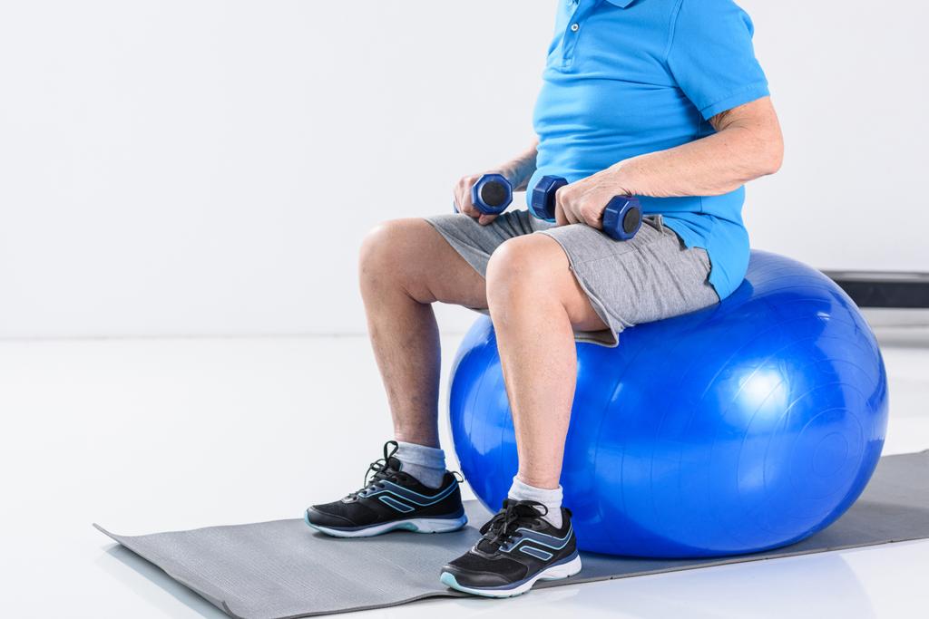 tiro recortado de homem sênior com halteres sentados na bola de fitness em fundo cinza
 - Foto, Imagem