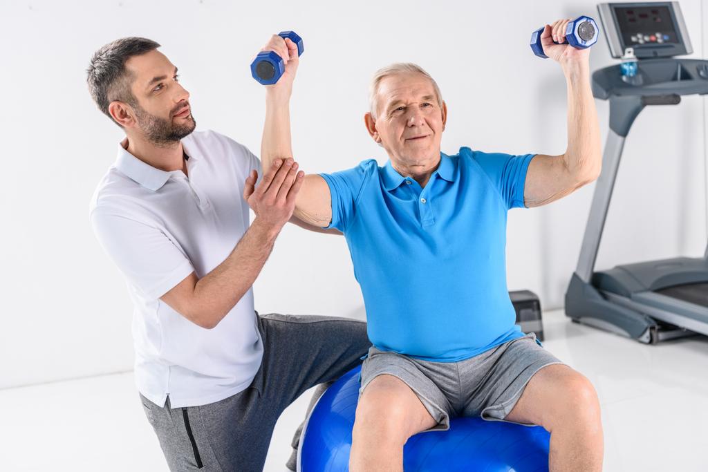 rehabilitation therapist assisting senior man exercising with dumbbells on fitness ball on grey backdrop - Photo, Image