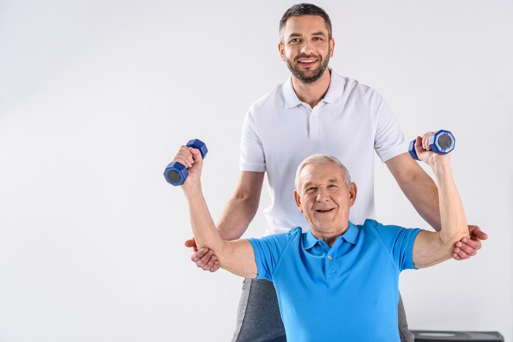 portrait of smiling rehabilitation therapist assisting senior man exercising with dumbbells isolated on grey - Photo, Image