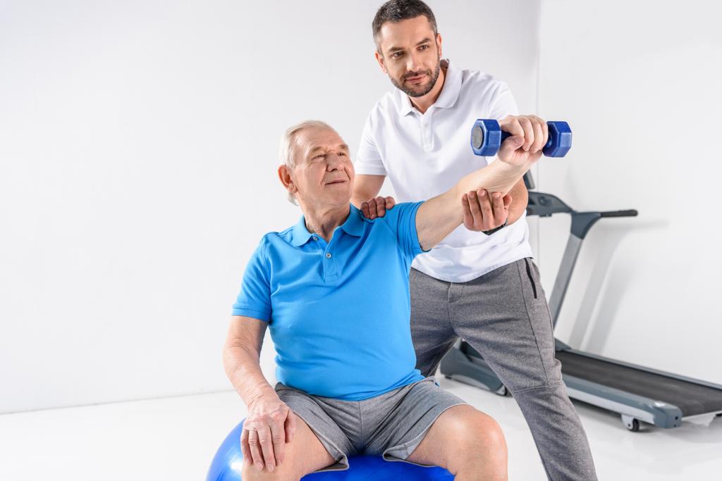 portrét rehabilitační pracovník pomáhat starší muž cvičení se činka na šedém pozadí - Fotografie, Obrázek