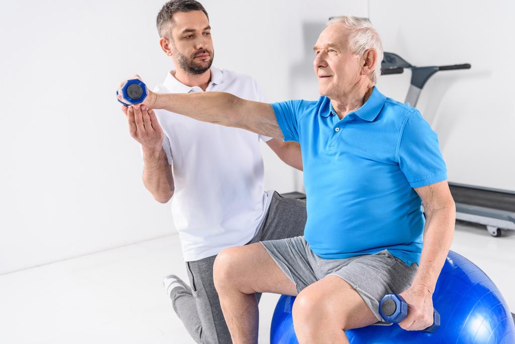rehabilitacji terapeuta pomoc starszy człowiek, ćwiczenia z hantlami na siłowni - Zdjęcie, obraz