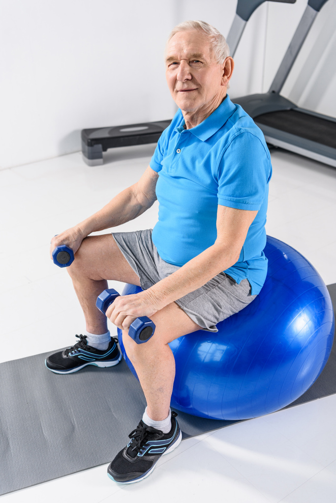 hoge hoekmening van senior man glimlachend met halters zittend op de fitness bal op grijze achtergrond - Foto, afbeelding