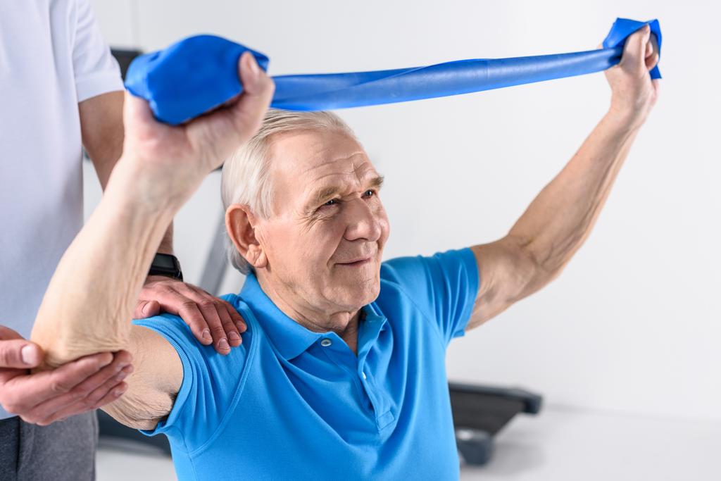 Oříznout záběr rehabilitační pracovník pomáhat starší muž cvičení s gumovou páskou - Fotografie, Obrázek