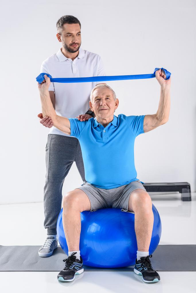 реабилитационный терапевт, помогающий старшему мужчине тренироваться с резиновой лентой на фитнес-мяч
 - Фото, изображение