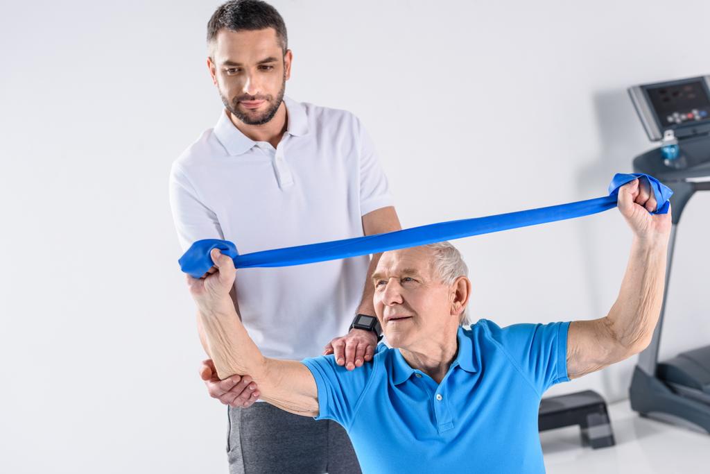 rehabilitation therapist assisting senior man exercising with rubber tape - Photo, Image