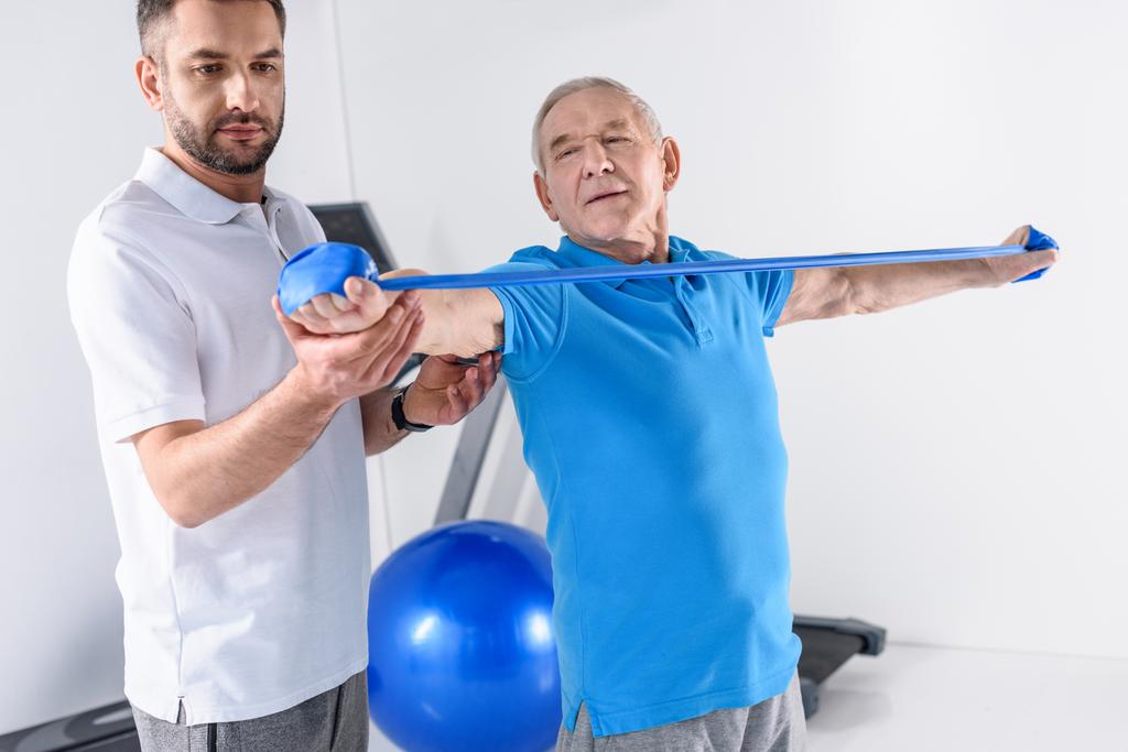 retrato del terapeuta de rehabilitación que ayuda al hombre mayor a hacer ejercicio con cinta de goma
 - Foto, imagen