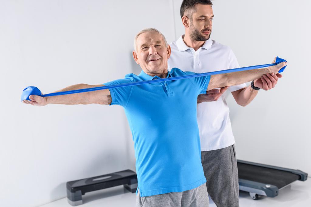 terapeuta de rehabilitación que ayuda a hombre mayor sonriente a hacer ejercicio con cinta de goma
 - Foto, Imagen