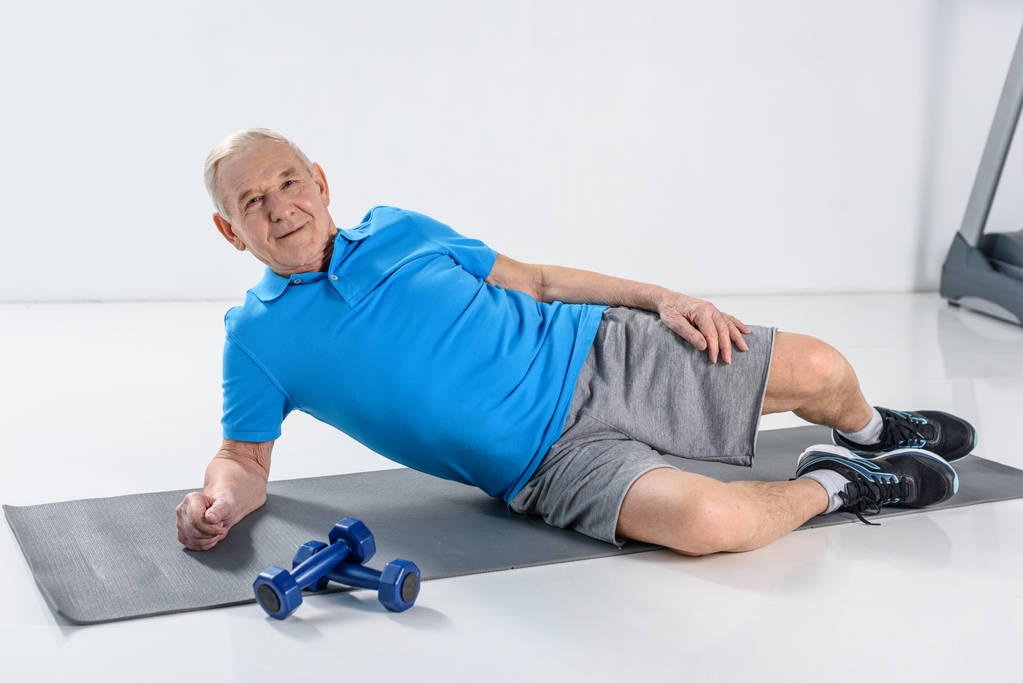 smiling senior man resting on mat with dumbbells near by on grey backdrop - Photo, Image