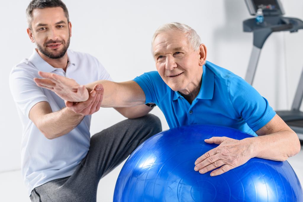 selective focus of rehabilitation therapist assisting senior man exercising on fitness ball - Photo, Image