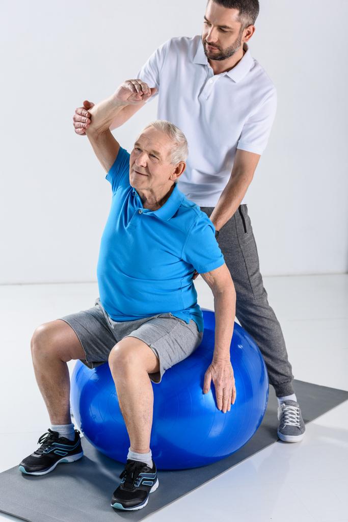 rehabilitation therapist assisting senior man exercising on fitness ball on grey backdrop - Photo, Image