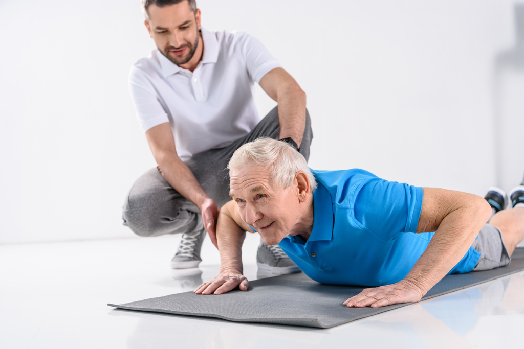 rehabilitacji terapeuta pomaga starszy człowiek ćwiczenia na macie na szarym tle - Zdjęcie, obraz