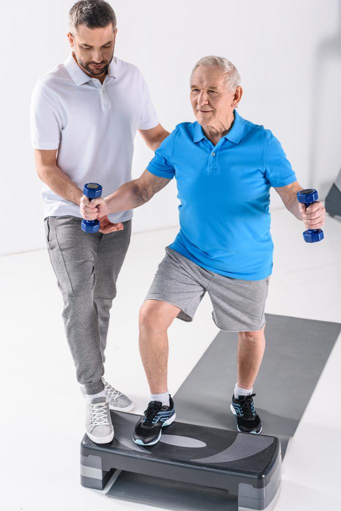 реабилитационный терапевт, помогающий старшему мужчине тренироваться с гантелями на сером фоне
 - Фото, изображение
