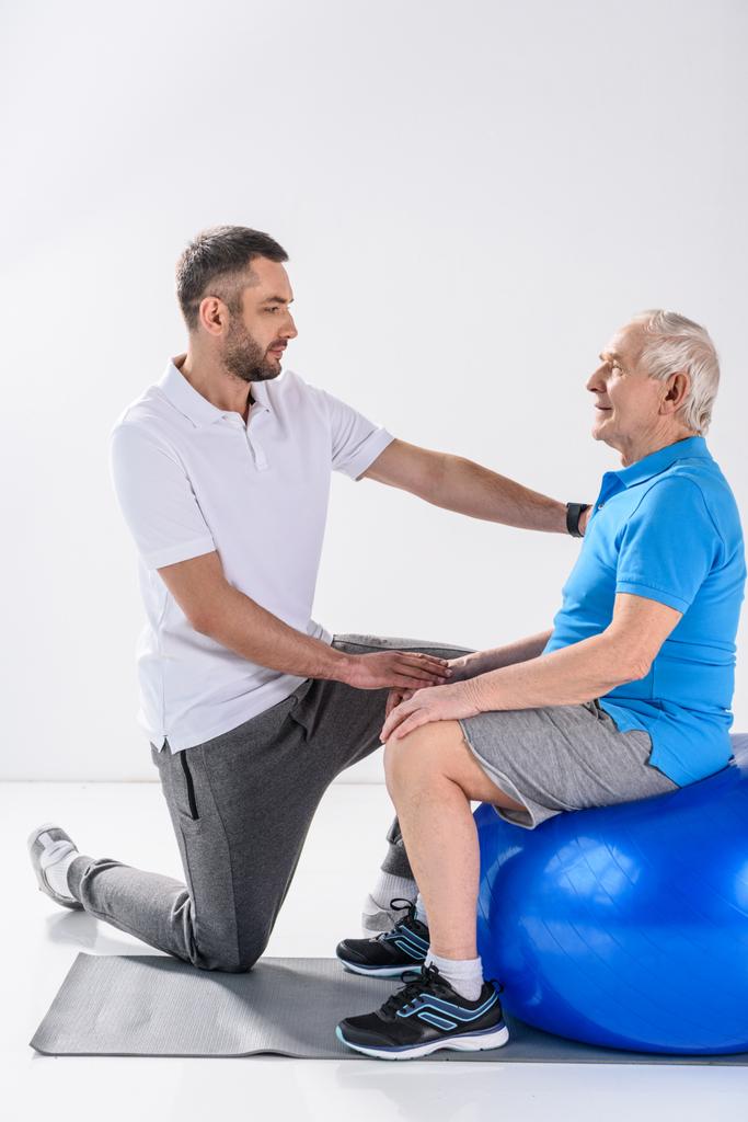вид збоку реабілітаційного терапевта, який допомагає старшому чоловікові тренуватися на фітнес-кулі на сірому тлі
 - Фото, зображення