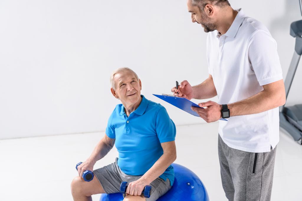 revalidatie therapeut met Kladblok bijstaan van senior man trainen met halters op grijze achtergrond - Foto, afbeelding