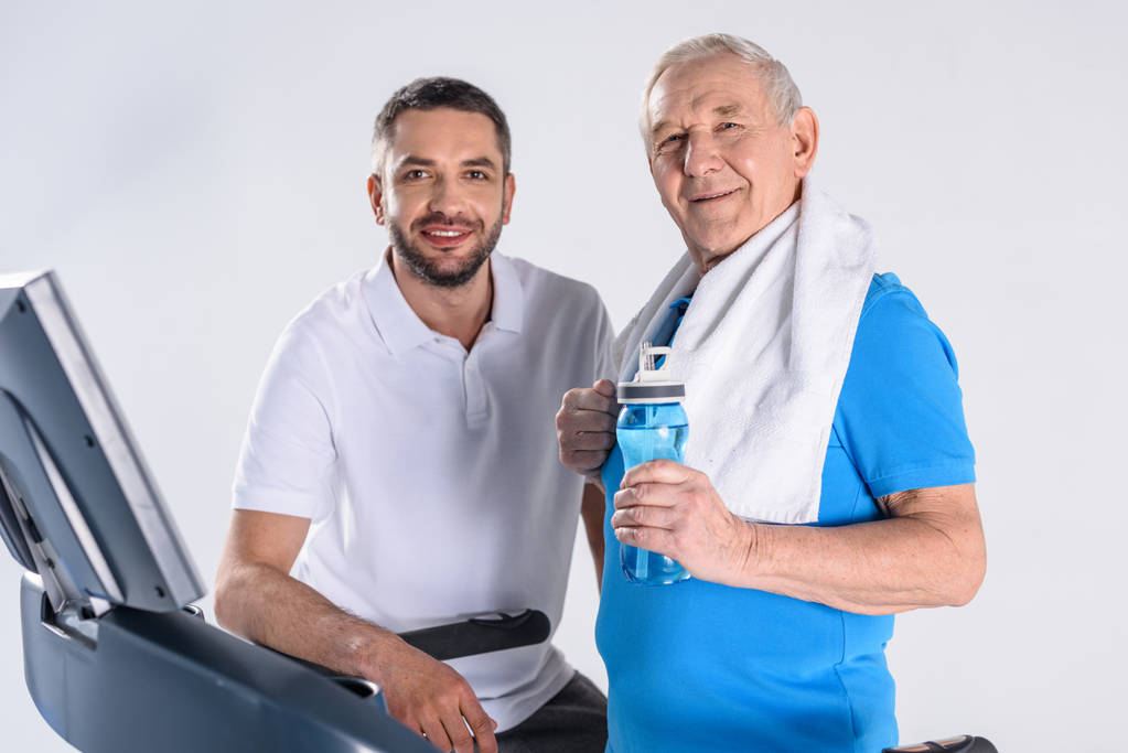 портрет улыбающегося реабилитационного терапевта и пожилого человека со спортивной бутылкой воды на беговой дорожке, изолированной на сером
 - Фото, изображение