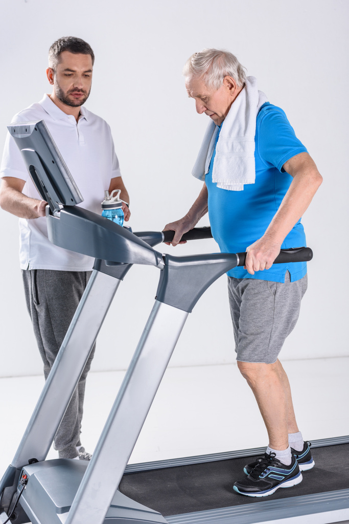 rehabilitation therapist assisting senior man with towel exercising on treadmill isolated on grey - Photo, Image