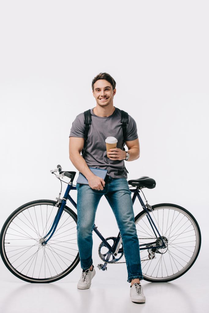 улыбающийся студент держит кофе, чтобы пойти и опираясь на велосипед на белом
 - Фото, изображение