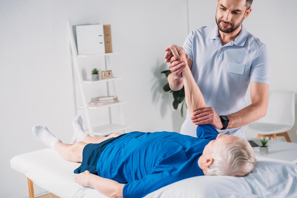 зосереджений фізіотерапевт робить масаж старшому чоловікові на масажному столі
 - Фото, зображення