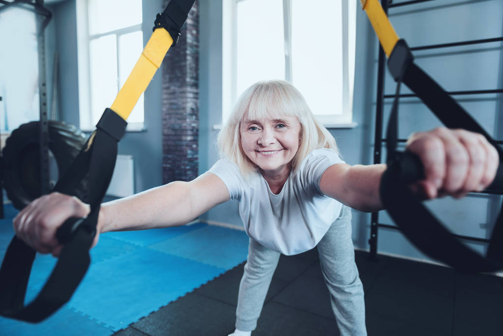 Mujer madura positiva sonriendo en la cámara mientras hace ejercicio en el gimnasio
 - Foto, imagen