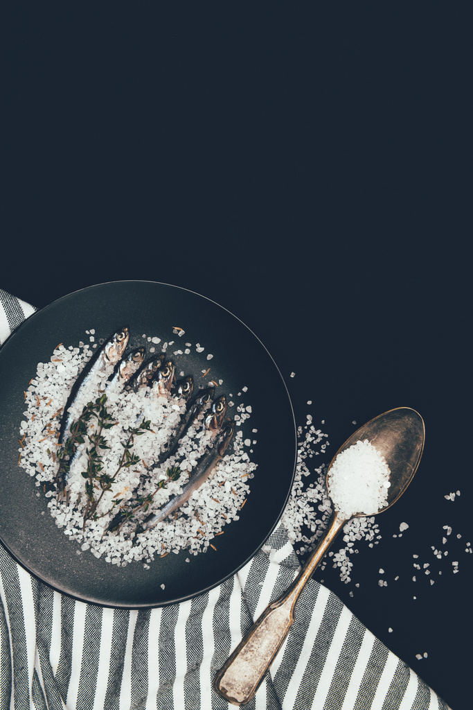 Frying pan with salted pile of fish, rosemary , spoon and towel isolated on black  - Photo, Image
