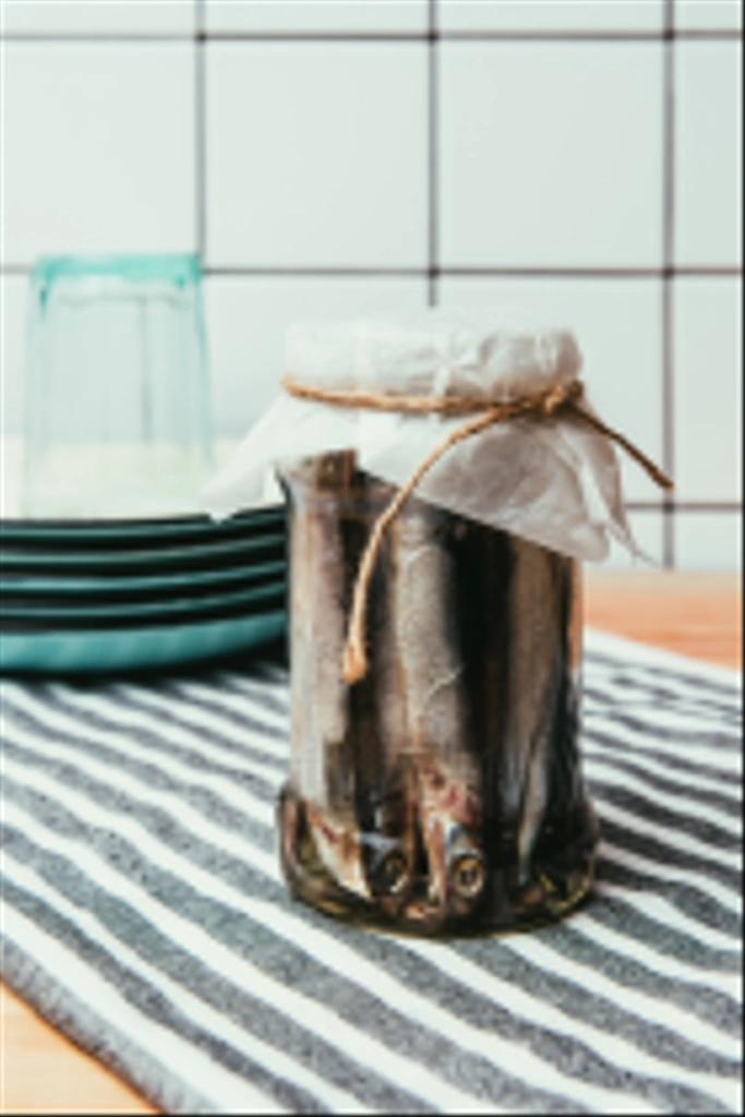 Купка солоної риби в банці, загорнута ниткою на рушник з тарілками і склом позаду
  - Фото, зображення