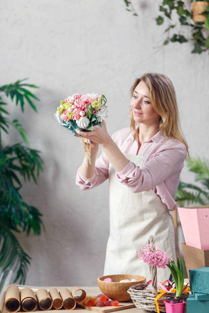 Imagem de florista segurando buquê em mãos à mesa com marmelada, marshmallow, caixas, papel
 - Foto, Imagem