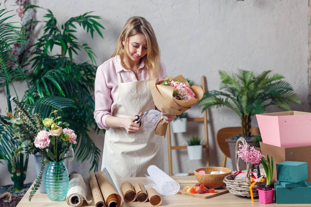 Photo of blonde florist in apron with bouquet with kraft paper on table with paper, marmalade - Photo, Image