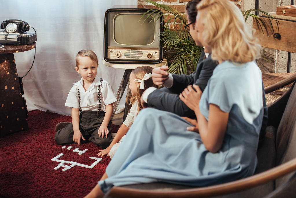 ouders op de bank zitten en kijken naar schattige kinderen spelen thuis, 50s stijl   - Foto, afbeelding