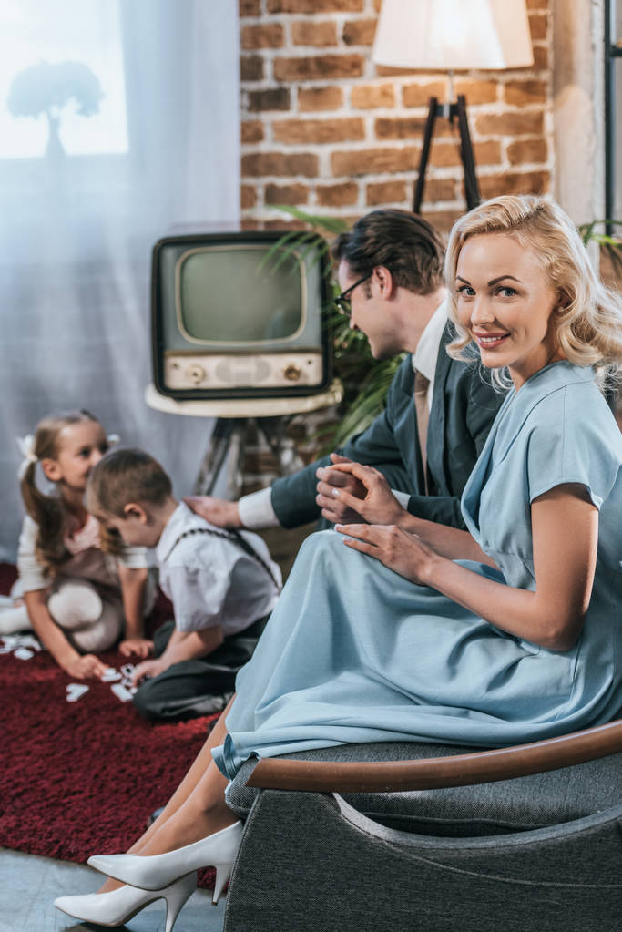 feliz mulher de estilo dos anos 50 sorrindo para a câmera enquanto sentado no sofá com marido e filhos brincando com telhas de dominó
 - Foto, Imagem
