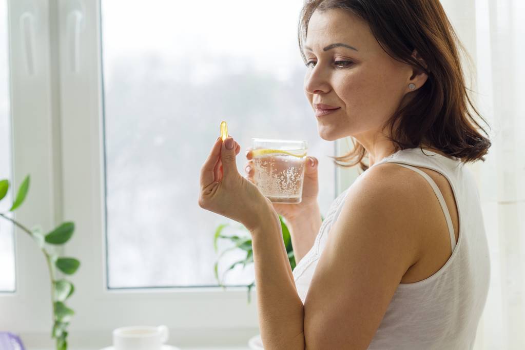 Žena má prášek s omega-3 a drží sklenici čerstvé vody s citronem. Obrázek domu, ráno u okna. - Fotografie, Obrázek