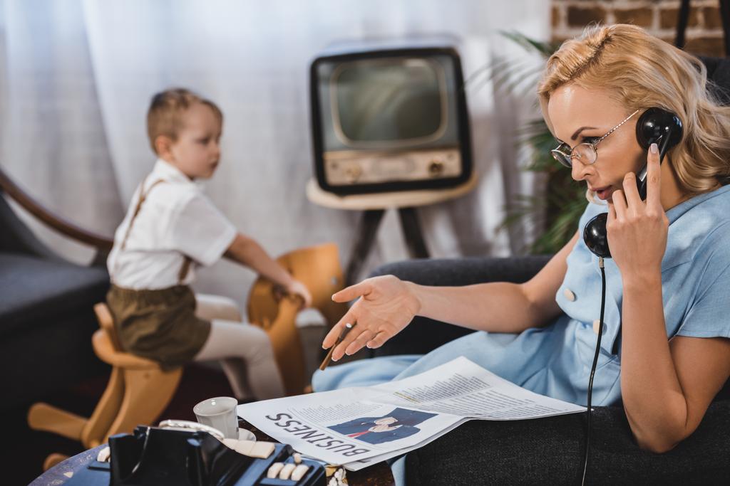 woman in eyeglasses reading business newspaper and talking by vintage telephone while little son sitting on rocking horse behind, 50s style family - Photo, Image