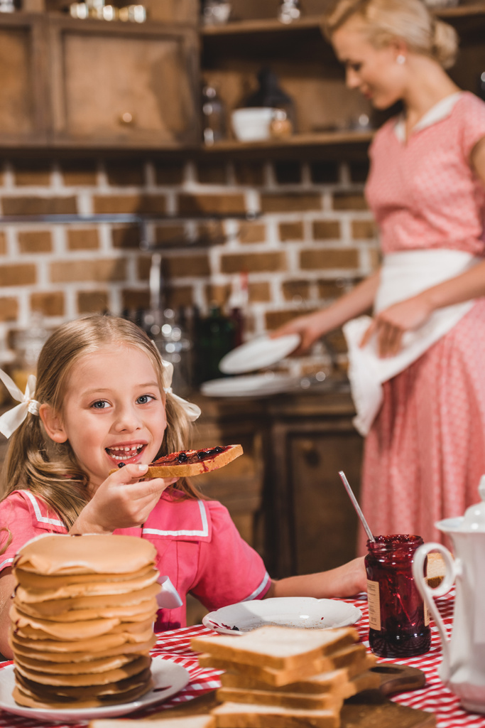 Rozkošná holčička jíst toasty s džemem a usmívá se na kameru, zatímco matka myje nádobí za styl roku 1950 - Fotografie, Obrázek