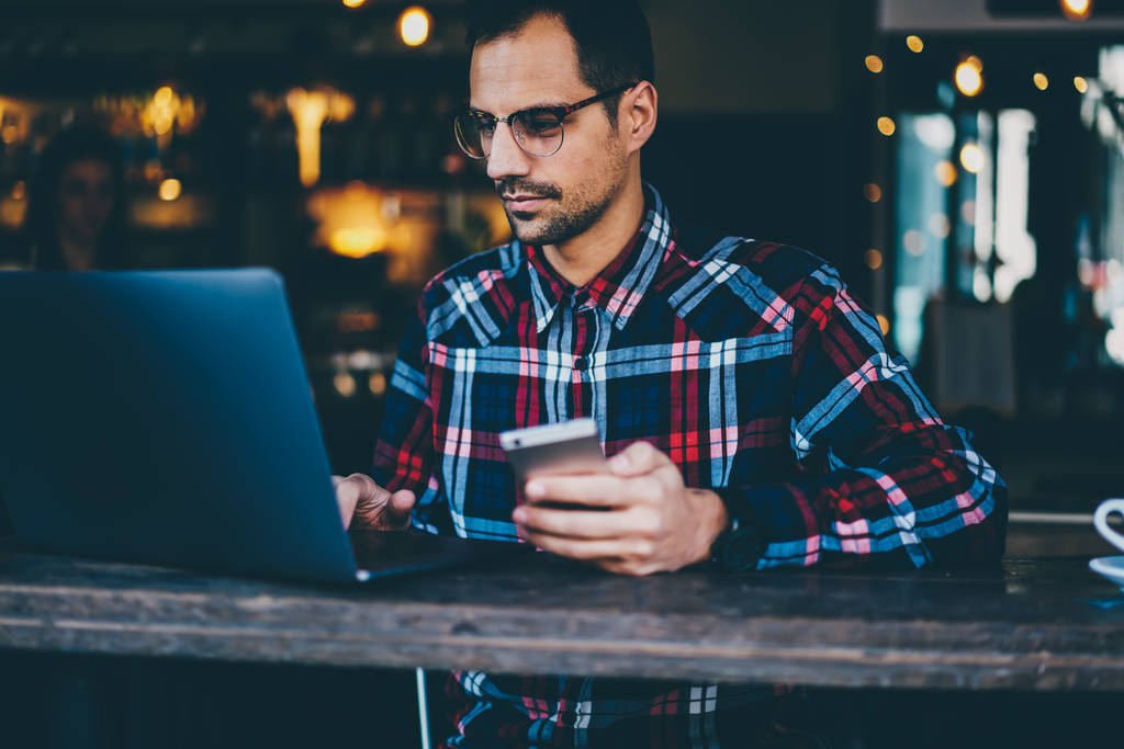 Профессиональный бородатый программист в очках, обновляющий программное обеспечение на современном ноутбуке, подключенном к беспроводному интернету, держа в руке цифровой смартфон, сидя в салоне кофейни
 - Фото, изображение