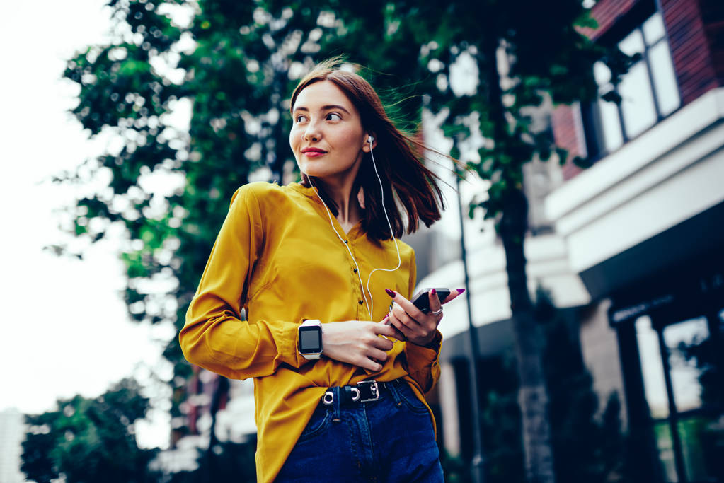 Young charming female blogger dressed in fashionable clothes enjoying recreation time with favorite music via earphones.Attractive redhead woman listening to playlist during summer walk outdoors - Photo, Image