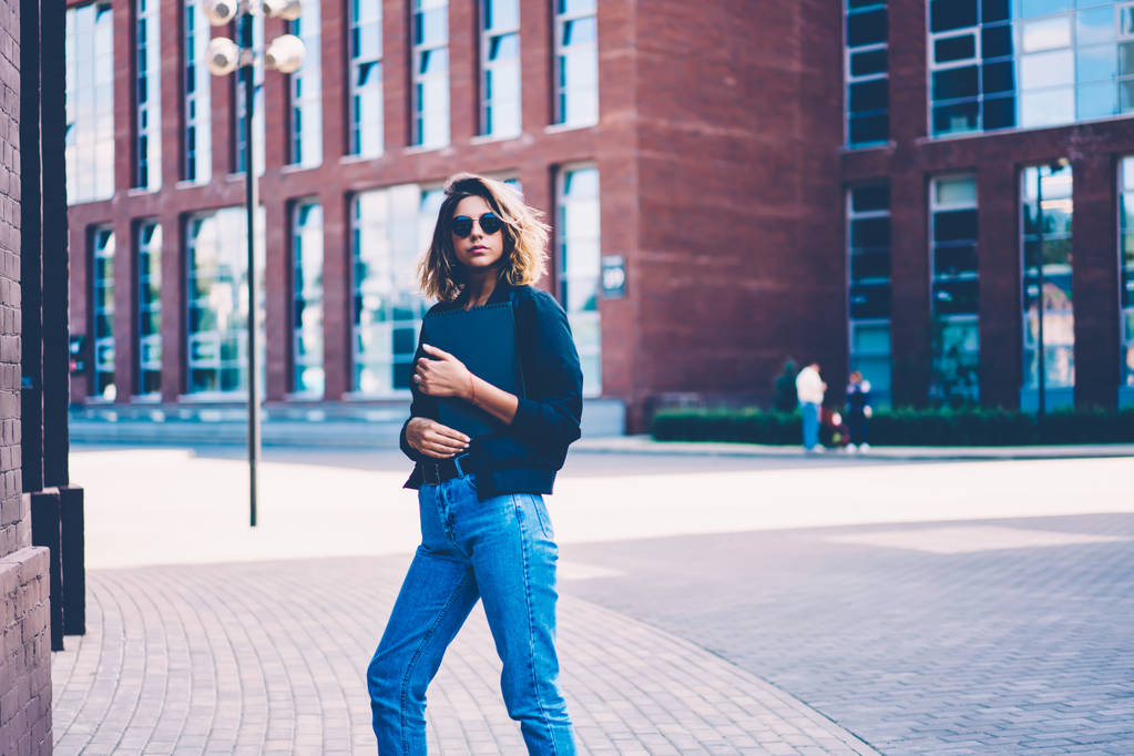 Jolie étudiante en lunettes de soleil marchant avec un bloc-notes dans la cour de l'université se dépêchant de se rencontrer, belle fille hipster vêtue de vêtements à la mode tenant dossier debout sur la zone de publicité urbaine
 - Photo, image