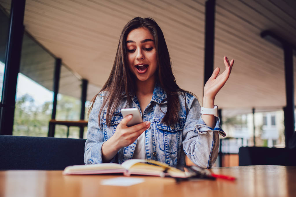 Emotionele hipster meisje kreeg geschokt nieuws op moderne smartphone. Verrast vrouw leest sms-bericht op digitale telefoon tijdens het lezen van interessante boek in coworking space - Foto, afbeelding
