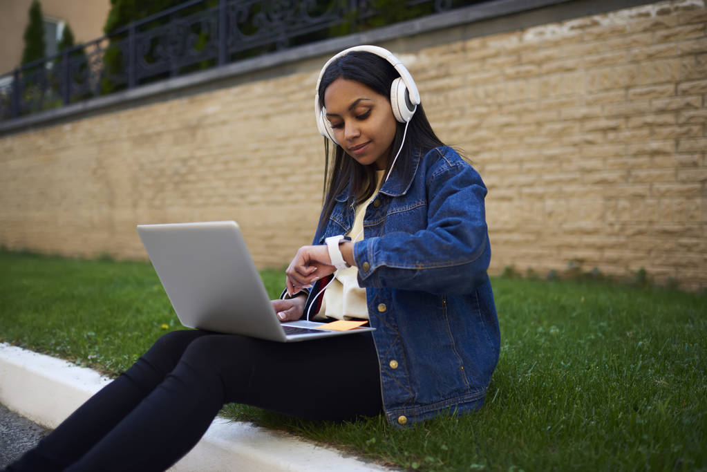 Estudante inteligente preparando lição de casa no campus universitário gerenciando o tempo para terminar antes do prazo, menina afro-americana em fones de ouvido ouvindo música da lista de reprodução do laptop verificando o tempo em brea
 - Foto, Imagem