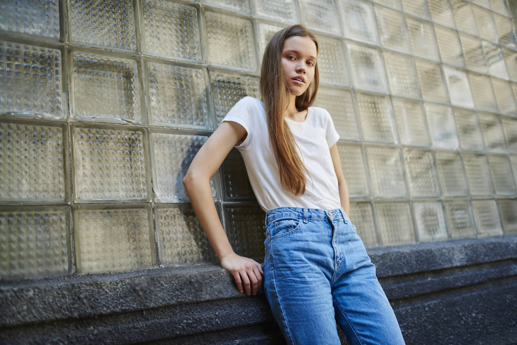 Portrét atraktivní mladá žena v módní oblečení, stojící na zdi městské skla, krásné teen model v bílé tričko s kopie prostoru pro vaše obchodní název nebo popisek pózuje pro camer - Fotografie, Obrázek