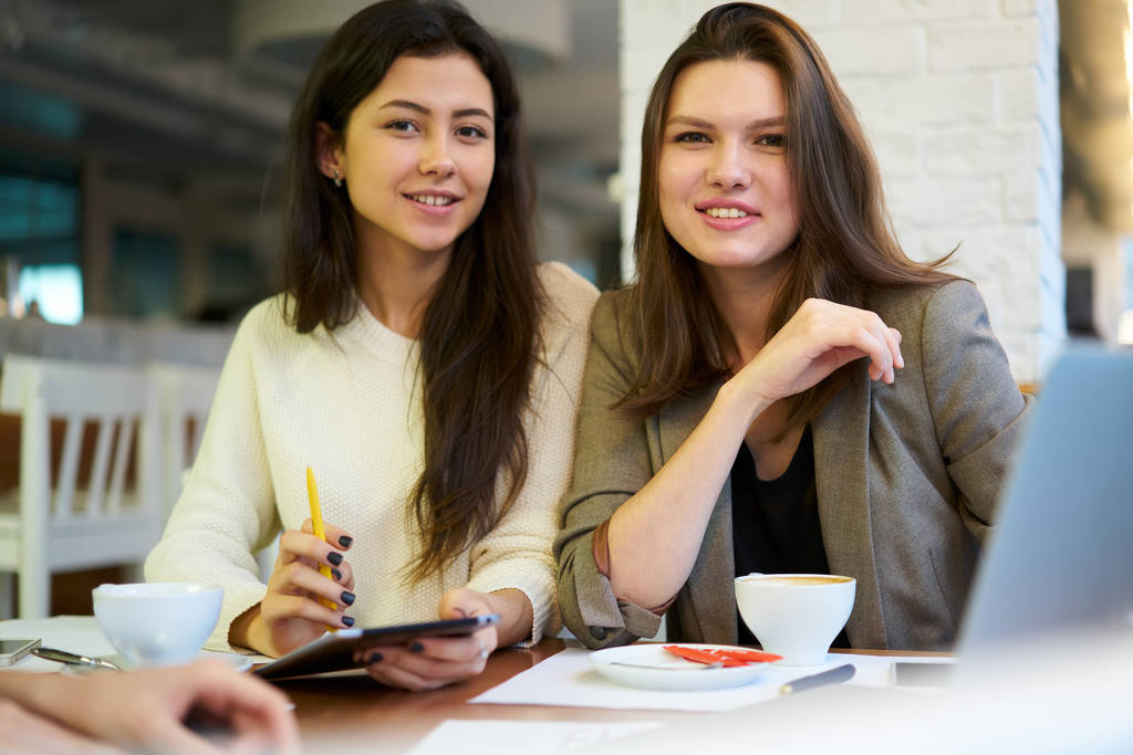 Портрет молодих процвітаючих жінок-колег задоволених результатом продуктивної роботи та співпраці над проектом, посміхнені ділові партнери дивляться на камеру, сидячи за столом у кафе спілкуючись
 - Фото, зображення