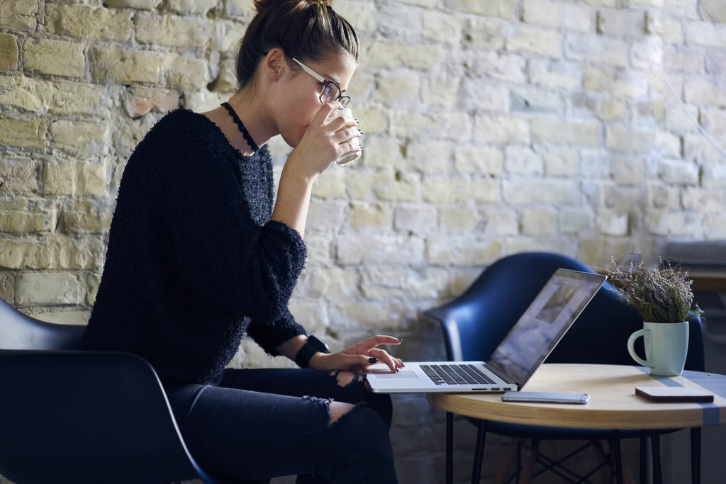 Συμπυκνωμένο έξυπνος μαθητής σε γυαλιά οράσεως και μοντέρνα ρούχα που εργάζονται στο φορητό υπολογιστή για την αναζήτηση πληροφοριών στο διαδίκτυο μαθημάτων πίνοντας νόστιμο καφέ κάθεται σε coworking χώρο - Φωτογραφία, εικόνα