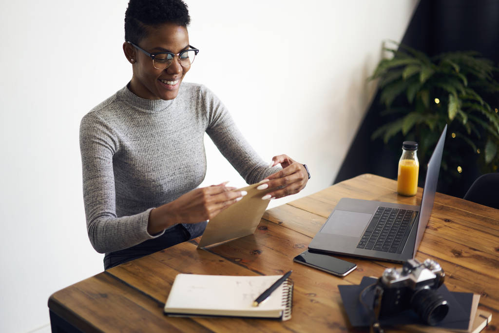 Fotógrafa afro-americana alegre empacotando fotos em envelope para enviar para postar ganhar dinheiro usando a tecnologia, sorrindo gerente em óculos satisfeito com a obtenção de papéis de negócios no correio
 - Foto, Imagem