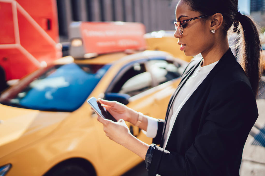 Mujer de negocios corporativa confiable que utiliza la aplicación de teléfono inteligente para llamar a taxi amarillo mientras está parado en el entorno urbano. Empresaria joven afroamericana que envía un mensaje por teléfono celular
 - Foto, Imagen