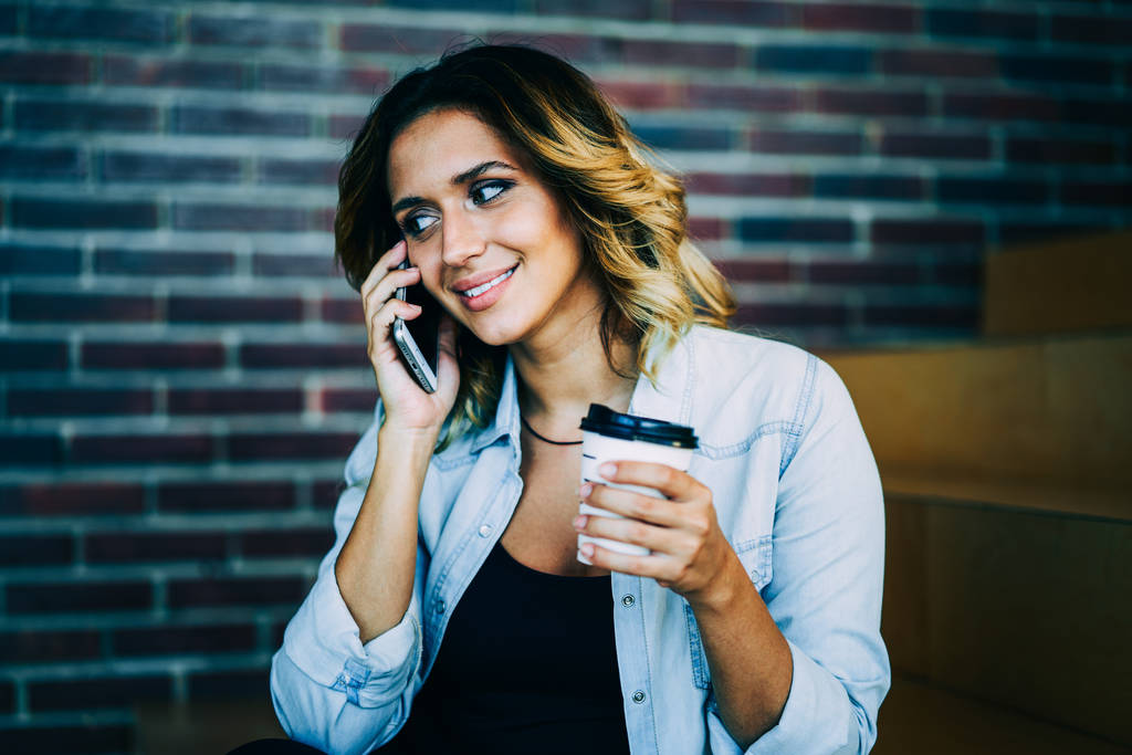 Mladá usmívající se dívka bederní komunikace s přáteli pomocí aplikace na chytrém telefonu připojený k Wi-Fi. Okouzlující pozitivní žena mluví o telefonu a drží aroma nápoje uvnitř - Fotografie, Obrázek
