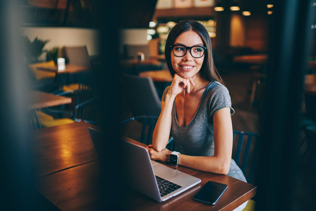 Atractiva freelancer femenina en gafas pensando en la idea para el proyecto de trabajo en el interior de la cafetería, mujer joven soñadora en gafas de actualización de software en netbook para hacer informe en línea para el trabajo remoto
 - Foto, imagen