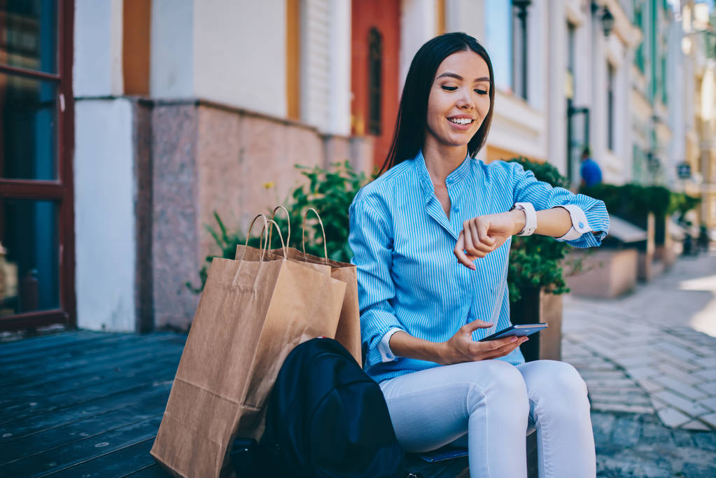 Beautiful smiling female person dressed in trendy clothing checking time on smartwatch while sitting and waiting for meeting outdoors.Gorgeous cheerful hipster girl spending free time at street - Photo, Image